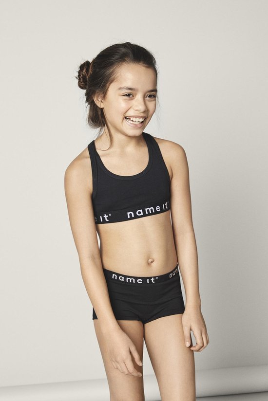 Lachend meisje met donker haar in een knot, draagt een zwart sporttopje en korte broek met 'name it' logo.