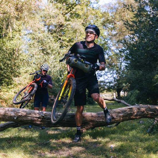 Man en vrouw met fietsen, klimmend over een omgevallen boom in een bosrijk gebied.