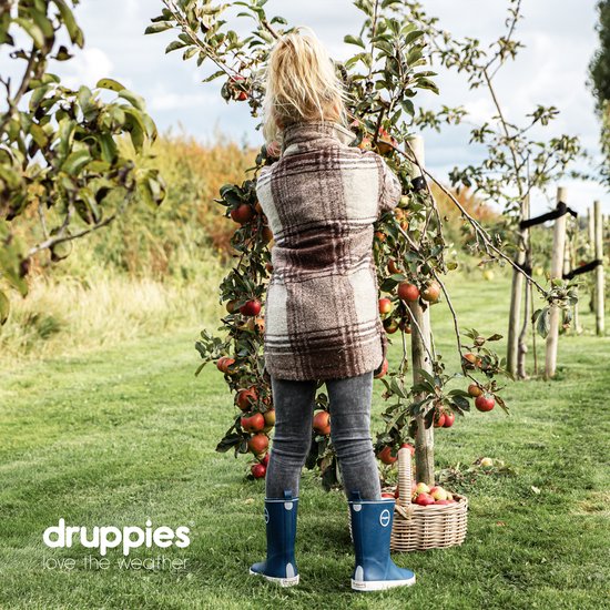 Meisje met blonde paardenstaart, staand in een boomgaard, draagt een geruite jas, grijze jeans en donkerblauwe rubberlaarzen.