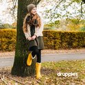 Meisje met blonde lokken, gele laarzen en winterjas, leunend tegen een boom.