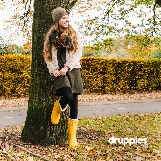 Meisje met blonde lokken, gele laarzen en winterjas, leunend tegen een boom.