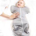 Een baby ligt in een grijze Swaddle UP slaapzak met witte strepen, met armen omhoog. Een hand rust op de buik.