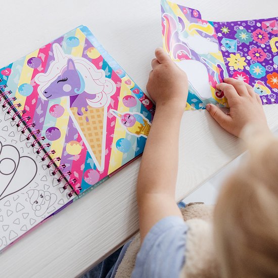 Une fille aux cheveux blonds en chignon, vue de dos, déplie une feuille d'autocollants Licorne violets, bleus et jaunes. Un livre ouvert avec une licorne sur un arc-en-ciel est à côté d'elle.