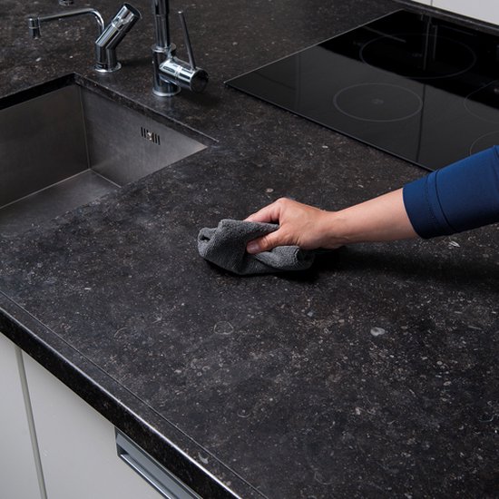 Hand met doek maakt een donker natuurstenen aanrechtblad schoon, met spoelbak en kraan.