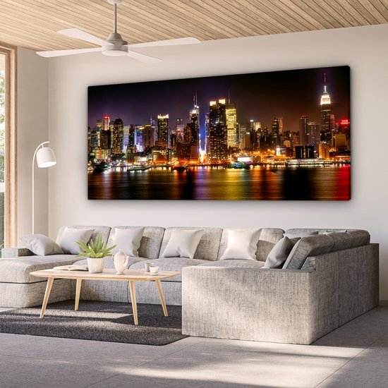 Un grand salon avec un canapé d'angle gris et une toile panoramique de la skyline nocturne de New York au mur.
