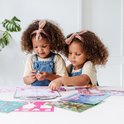 Deux jeunes filles aux cheveux bouclés et bandeaux roses, dessinant à une table avec du matériel Unicorn Academy.