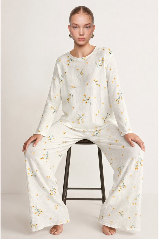 Model zittend op een kruk, gekleed in een ecru pyjamaset met een fijne gele bloemenprint en wijde broekspijpen.