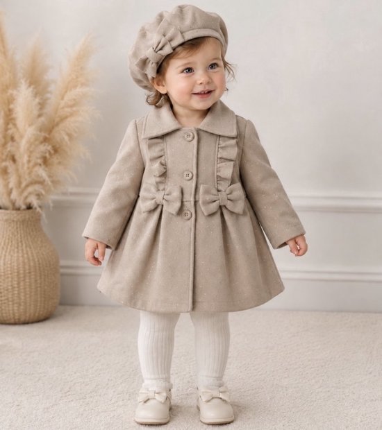 Meisje in een beige jas met ruches, strikken en bijpassende baret. Witte maillot en schoentjes.