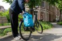 Persoon die een helderblauwe Voltano fietstas op een zwarte e-bike bevestigt in een groene buitenomgeving.