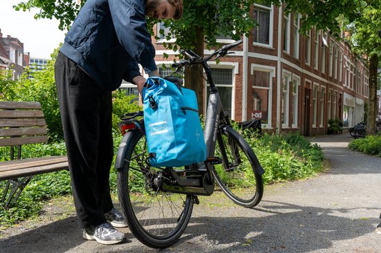 Persoon die een helderblauwe Voltano fietstas op een zwarte e-bike bevestigt in een groene buitenomgeving.