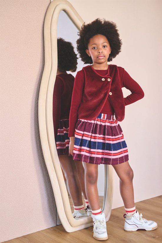 Model draagt een bordeauxrood faux fur gilet over een bordeauxrode top en een rok met veelkleurige strepen.