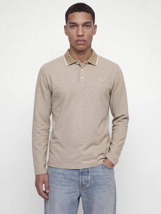 Model in beige poloshirt met lange mouwen, bruine kraag met witte bies en lichtblauwe jeans.