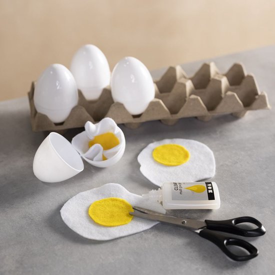 Witte plastic eieren, vilt-eieren met gele dooiers, schaar en een flesje Clear multi glue op een grijze ondergrond.