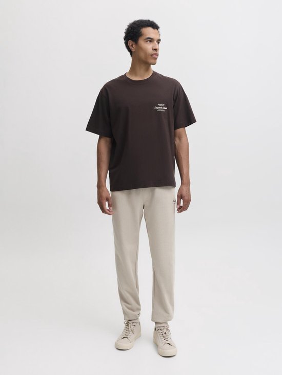 Man in bruin T-shirt met witte tekst op de borst en beige joggingbroek.