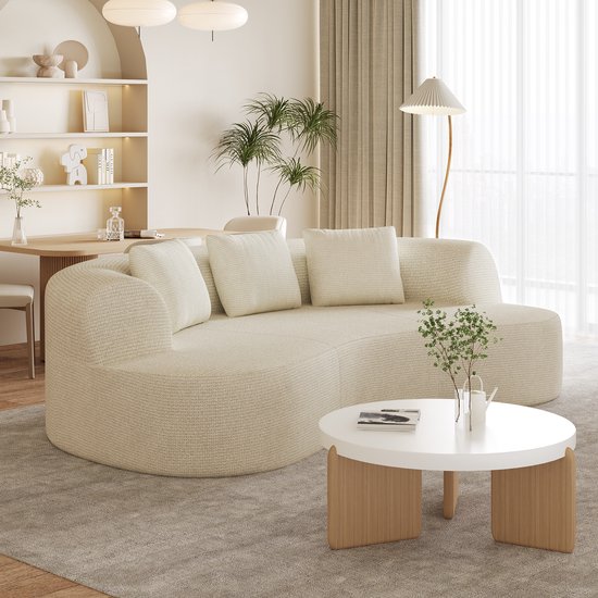 Crèmekleurige, gebogen bank met een textuur van chenille en vier dikke, losse kussens. Een ronde, witte salontafel met houten poten staat ervoor.