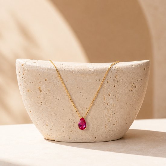 Gouden ketting met een rode robijnkleurige druppelvormige edelsteen, gepresenteerd op een betonnen sculptuur.