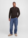 Model draagt bruine polo met lange mouwen, jeans en beige schoenen, hand in broekzak.