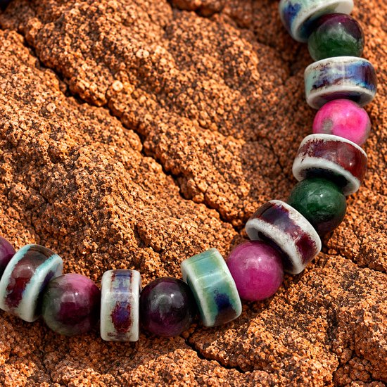 Close-up van een armband met ronde paarse, groene en roze stenen en geglazuurde keramische kralen met witte patronen.