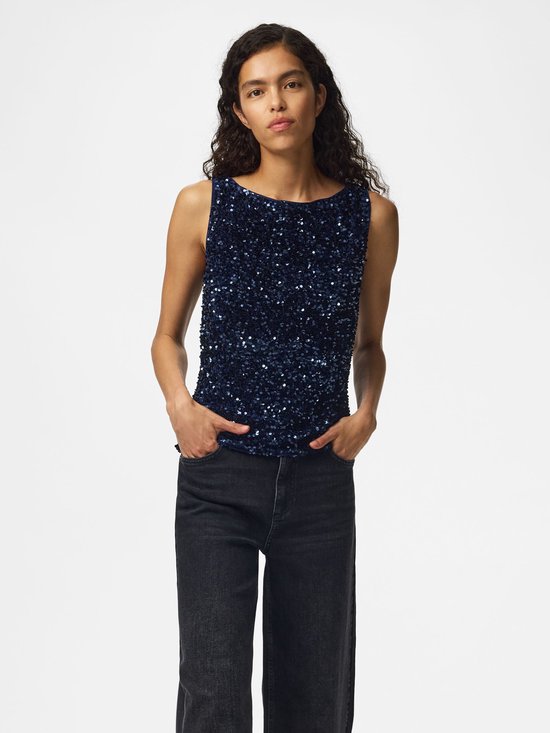 Een vrouw met lang krullend haar draagt een donkerblauw mouwloos topje met pailletten en een zwarte jeans.