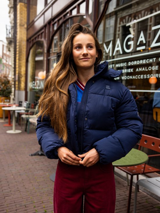 Vrouw met lang bruin haar, buiten in een donkerblauwe pufferjas, met rode broek, bij een etalage.