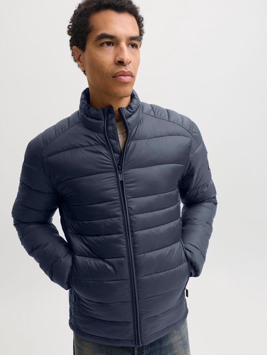 Model met handen in de zakken, draagt een donkerblauw pufferjack met rits en opstaande kraag.