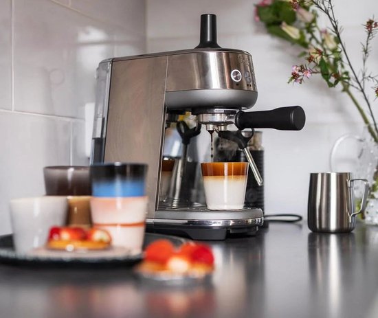 Zilverkleurige Sage espressomachine zet koffie in een tweekleurig glazen kopje tussen diverse kleurrijke aardewerken bekers.