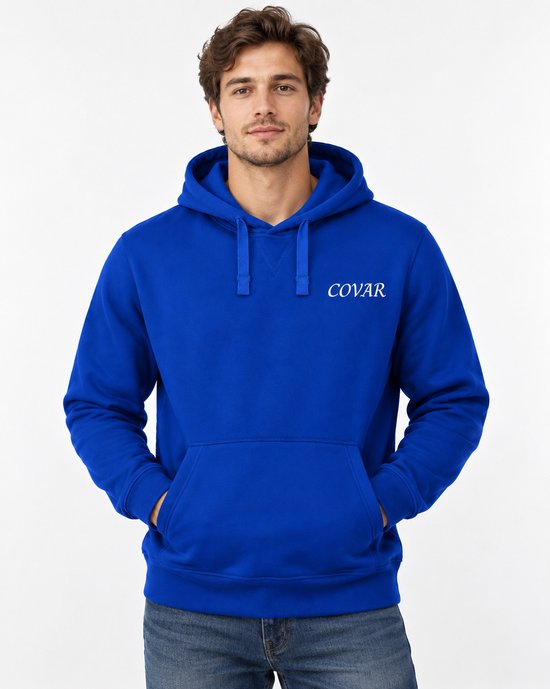 Man met bruin haar draagt een koningblauwe hoodie met 'COVAR' logo en jeans, handen in zakken.