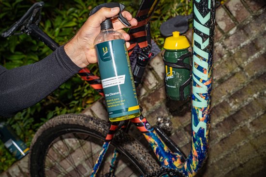Hand houdt U-Sport Bike Cleaner fles bij een kleurrijke fiets. Fles is groen met geel, fiets heeft patronen.