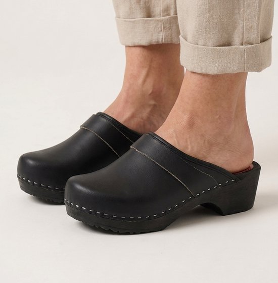 Zwarte Dina klompen met houten zool en witte stiksels, gedragen onder een beige linnen broek.