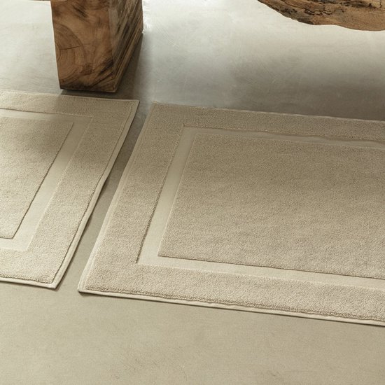 Een beige badmat met een dubbel, ingesneden randmotief op een betonnen vloer, deels zichtbaar is een andere mat en een houten meubel.