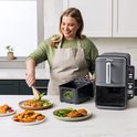 Vrouw met schort die kip en groenten serveert uit een zwarte Ninja heteluchtfriteuse.