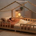 Enfant dormant dans un lit maison en bois naturel avec barreaux et guirlande lumineuse.