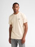 Man staat recht naar voren, draagt een crème T-shirt met opdruk en grijze jeans.