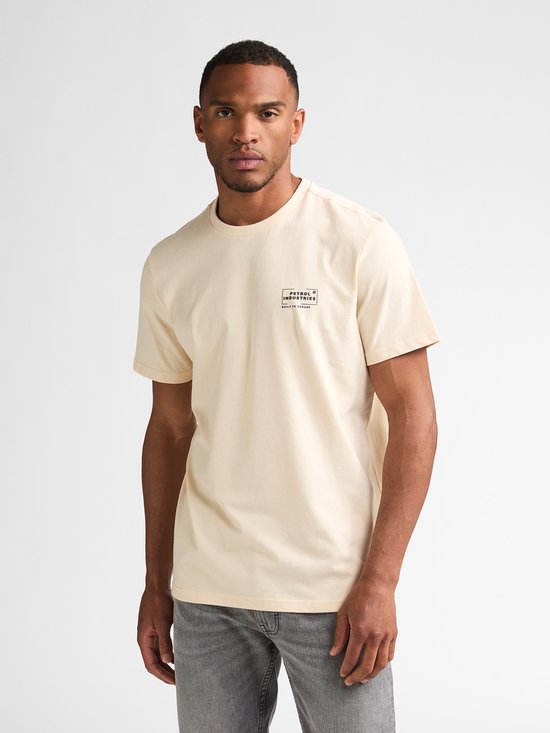 Man staat recht naar voren, draagt een crème T-shirt met opdruk en grijze jeans.