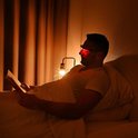 Man in bed leest boek, draagt bril met rood licht.