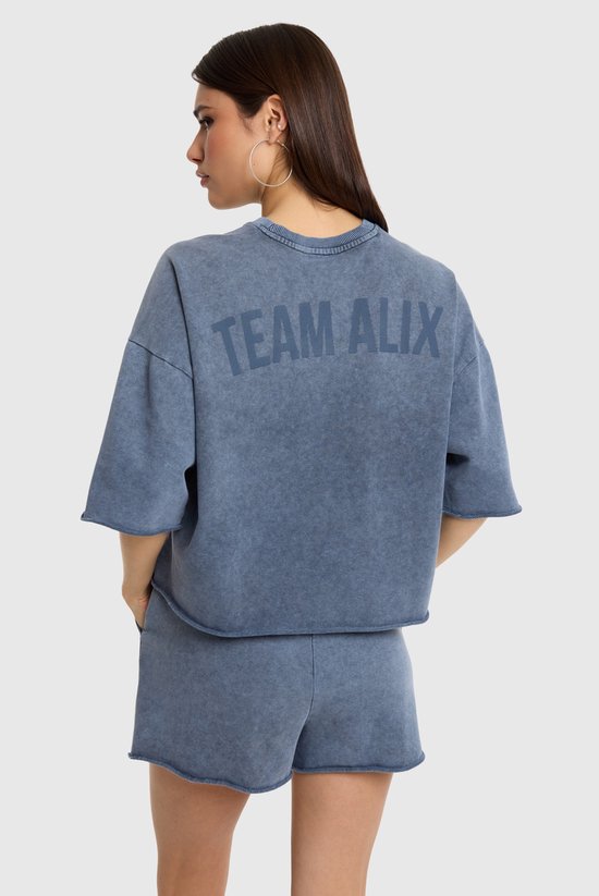 Model draagt een blauwe, geknipte top met korte mouwen en de tekst 'TEAM ALIX' op de rug.