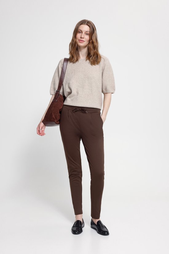 Model in een beige top en bruine broek met strikceintuur, steekzakken.