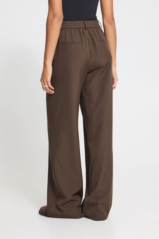 Model van achteren gezien, draagt een wijde bruine broek met elastische tailleband en een opgestikte achterzak.