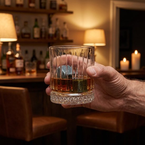 Een hand houdt een glas met whiskey en stenen vast in een baromgeving met gedimd licht.