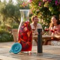 Glazen karaf met sangria en een turquoise deksel, naast een zwarte vacuümpomp op een houten tafel.