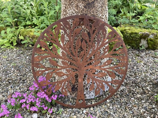Plaque murale ronde en métal rouillé avec un motif d'arbre de vie, devant un tronc d'arbre et des plantes.