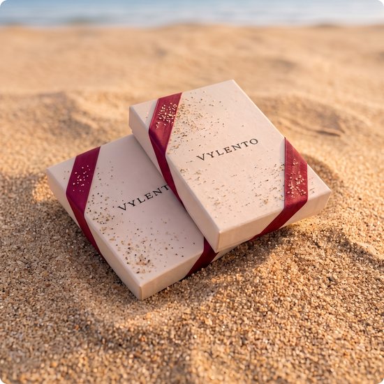 Twee geschenkdozen van Vylento met een rode strik en gouden glitter, liggend op het zand.
