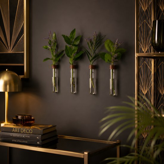 Vier glazen reageerbuisjes met planten en bladeren, opgehangen aan een donkere muur, naast boeken en een gouden tafellamp.
