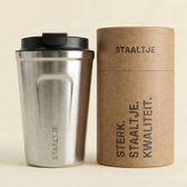 Zilverkleurige STAALTJE thermosbeker met zwarte deksel, naast de ronde verpakking met tekst.
