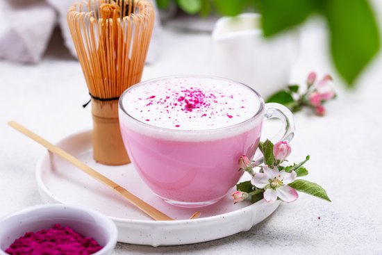 Een glas roze bietendrank met schuim, bestrooid met roze poeder, naast een bietenschepje en bloesems.