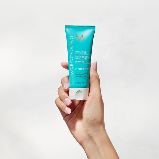 Hand die Moroccanoil Hydrating Styling Cream tube vasthoudt. Turquoise tube met oranje M-logo.