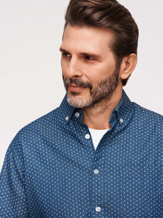 Close-up van man met baard, draagt een blauw overhemd met kleine witte stipjes over een wit t-shirt.