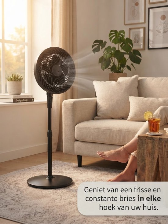 Zwarte staande ventilator in een woonkamer met een bank. Een vrouw zit op de grond naast de ventilator. Er staat een glas met drankje op tafel.