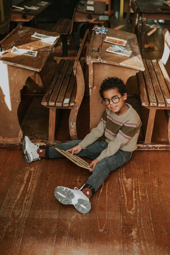 Jongen met krullend haar en bril, zittend op de grond in een oude schoolklas, draagt een gestreepte trui en grijze jeans.