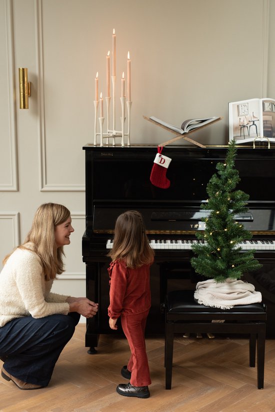Vrouw kijkt naar een kind in een roze houten kledingset met ruches, naast een piano.